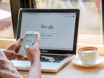 Person typing a search into mobile with a laptop open to Google in the background